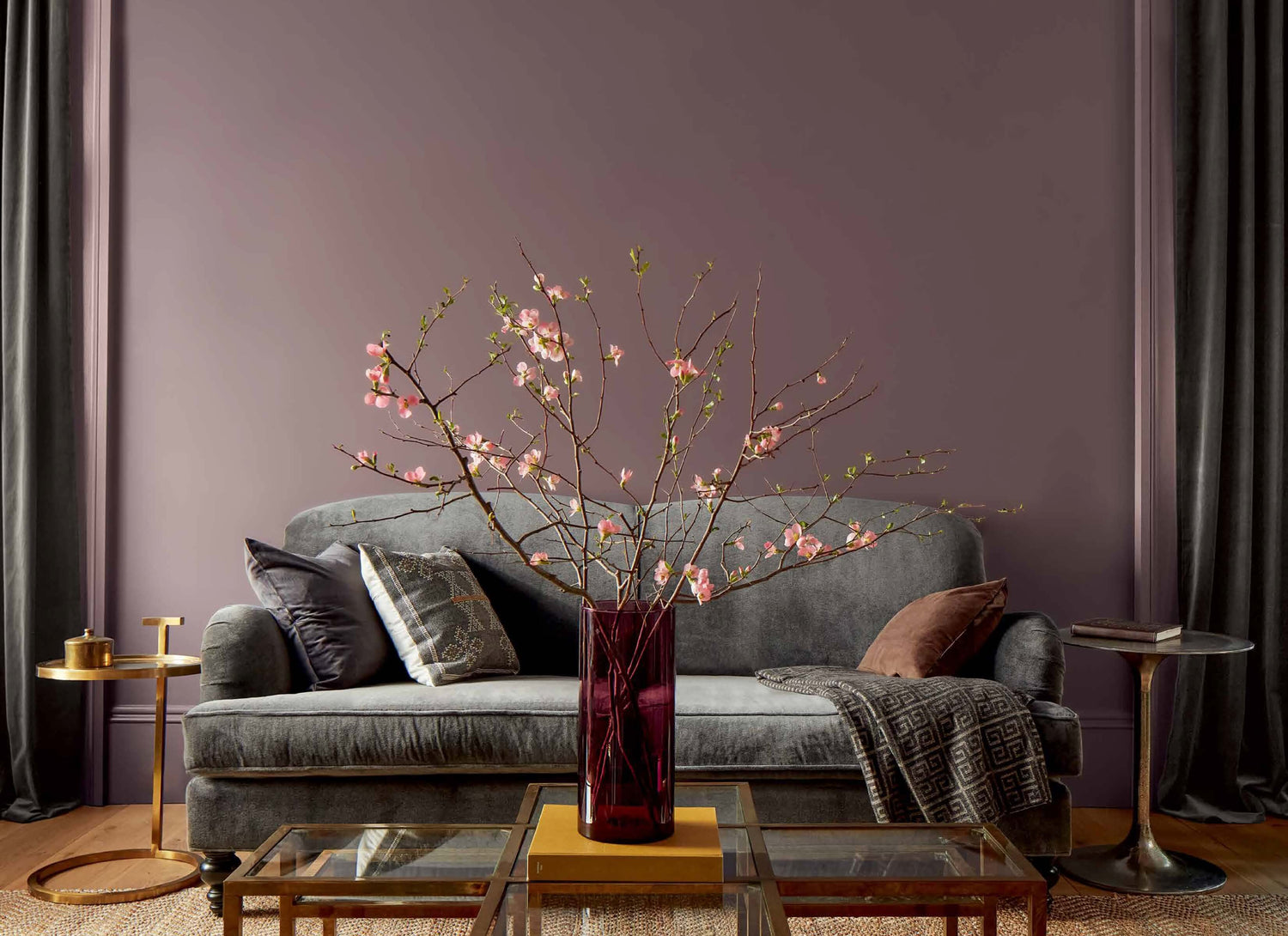A purple-painted living room features a grey couch, gold side tables, and a glass-topped gold coffee table topped with a red vase filled with pink flowers.