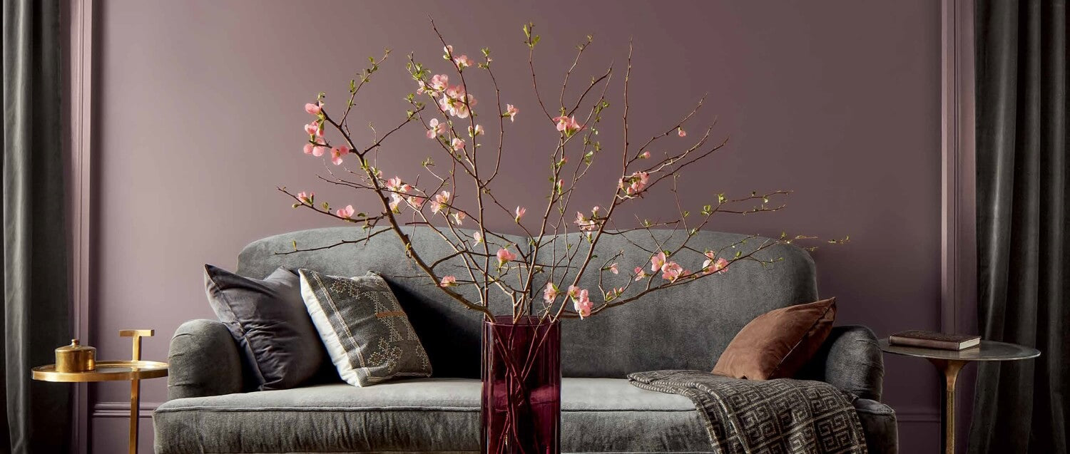 A purple-painted living room features a grey couch, gold side tables, and a glass-topped gold coffee table topped with a red vase filled with pink flowers.